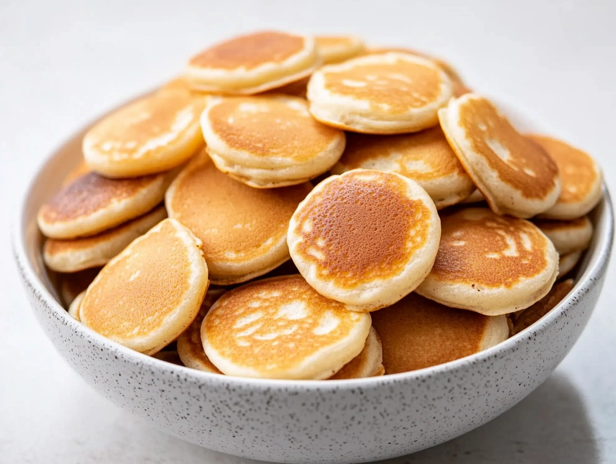 Neatly stacked mini pancakes on a white plate, with a ripe strawberry in the center and a soft mauve cloth draped in the background.