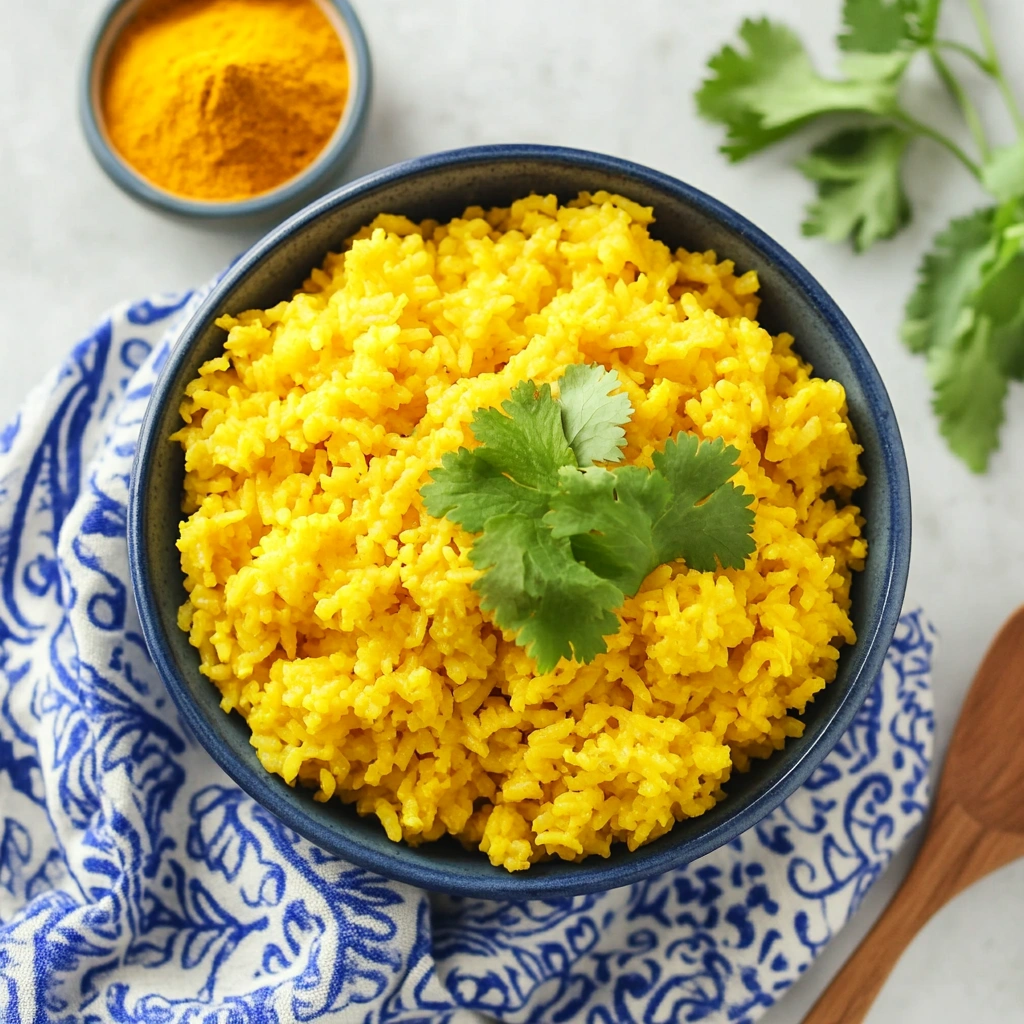 Bright yellow rice with herbs served in a white bowl, accompanied by a wooden spoon and fresh ingredients on a clean backdrop.