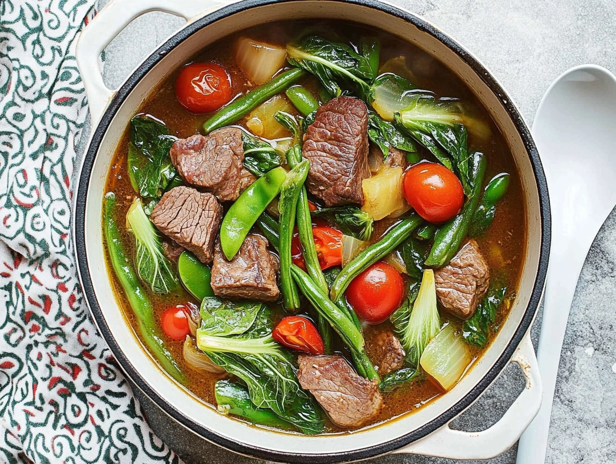 A top-down view of a pot filled with beef sinigang, featuring chunks of beef, green beans, bok choy, cherry tomatoes, and leafy greens in a steaming, sour broth.