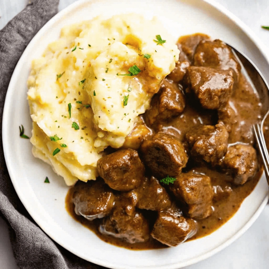 Juicy beef tips in gravy and fluffy mashed potatoes on a white plate.