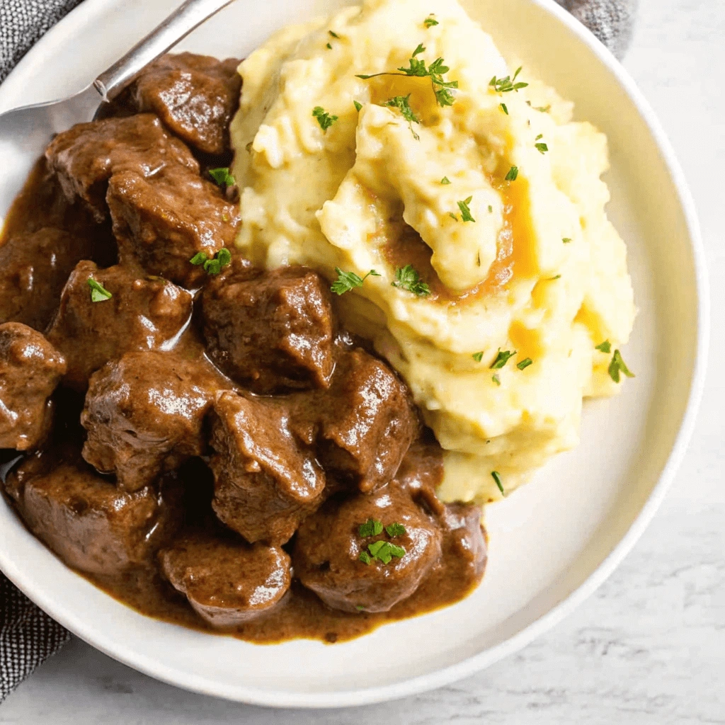 Close-up of tender beef tips with rich brown gravy served alongside creamy mashed potatoes garnished with parsley on a white plate.